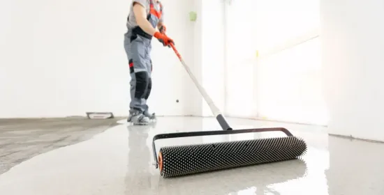 Worker using a floor roller to spread coating on a wet concrete floor in a bright room