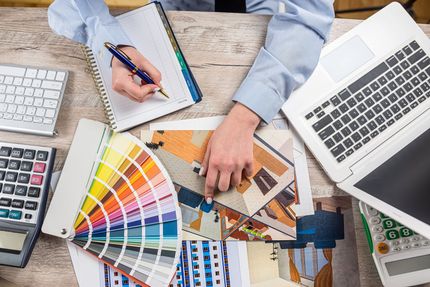 Designer reviewing color swatches and layout samples beside a laptop and calculator on a desk