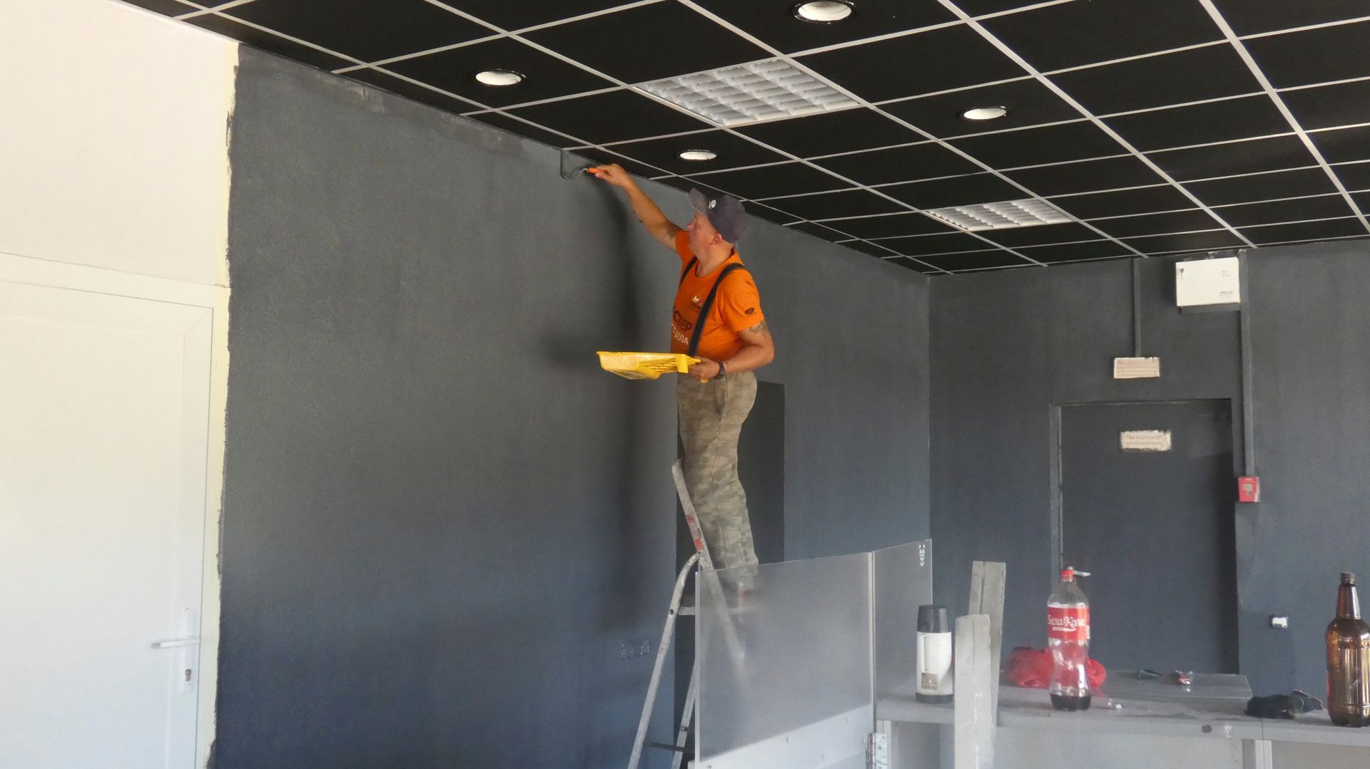 Worker in orange vest painting a dark wall from a ladder in a room under renovation