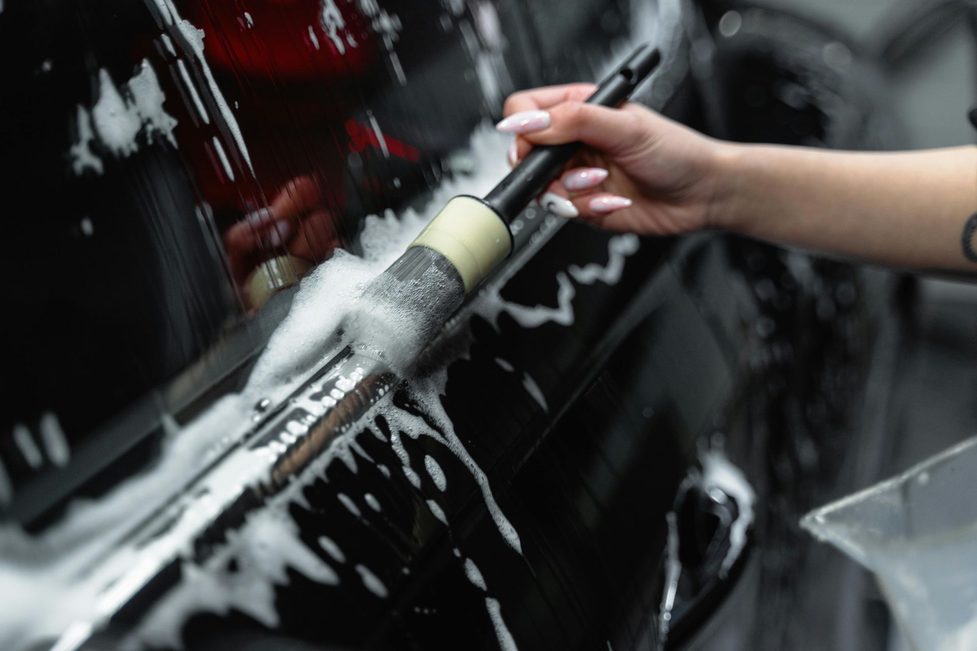 Hand with brush cleaning a black car, covered in white foam.