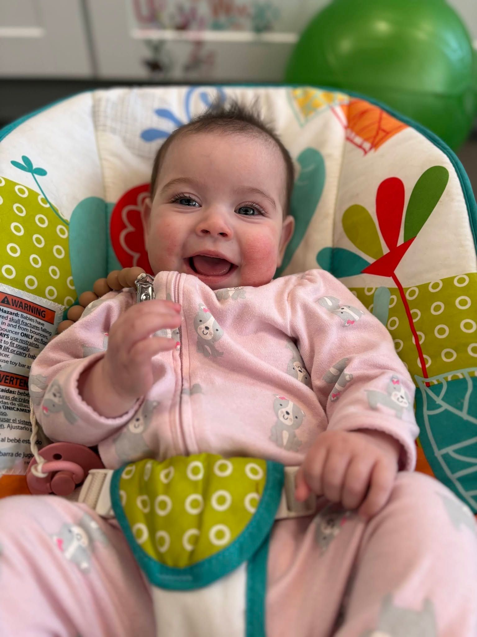 Smiling baby in a pink outfit sitting in a colorful patterned infant seat