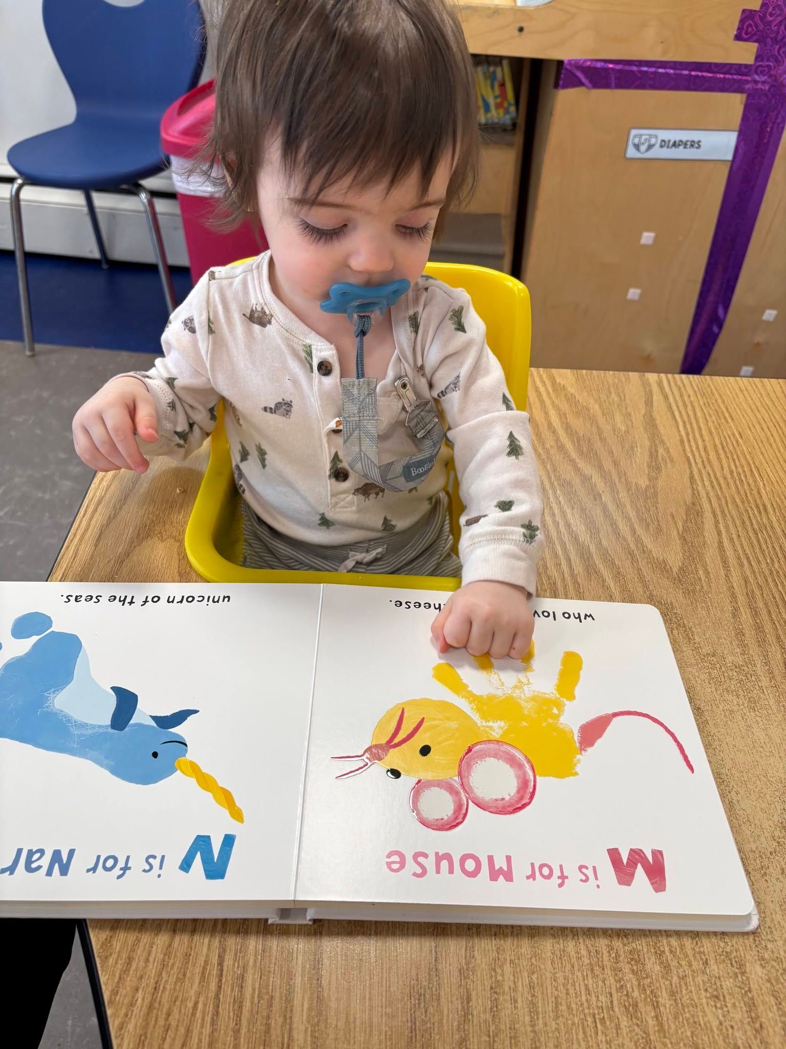 Toddler painting at a table with blue and yellow animal art pages, using a paintbrush and cup holder
