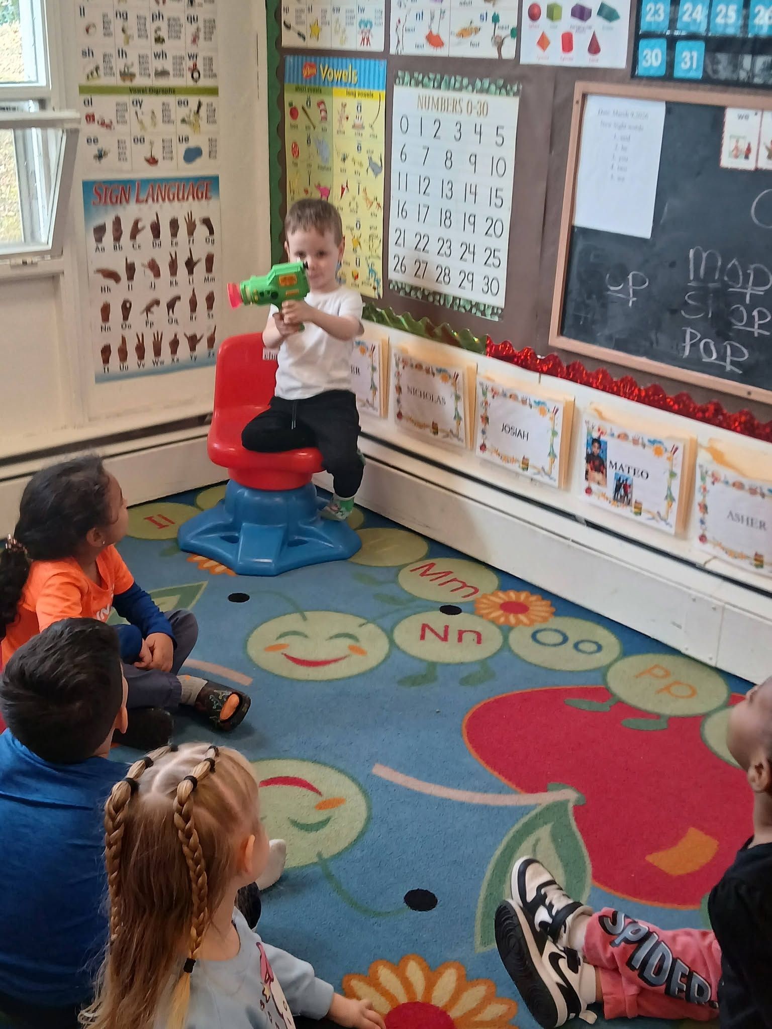 Child demonstrating a toy in a colorful classroom while classmates sit on a rug watching