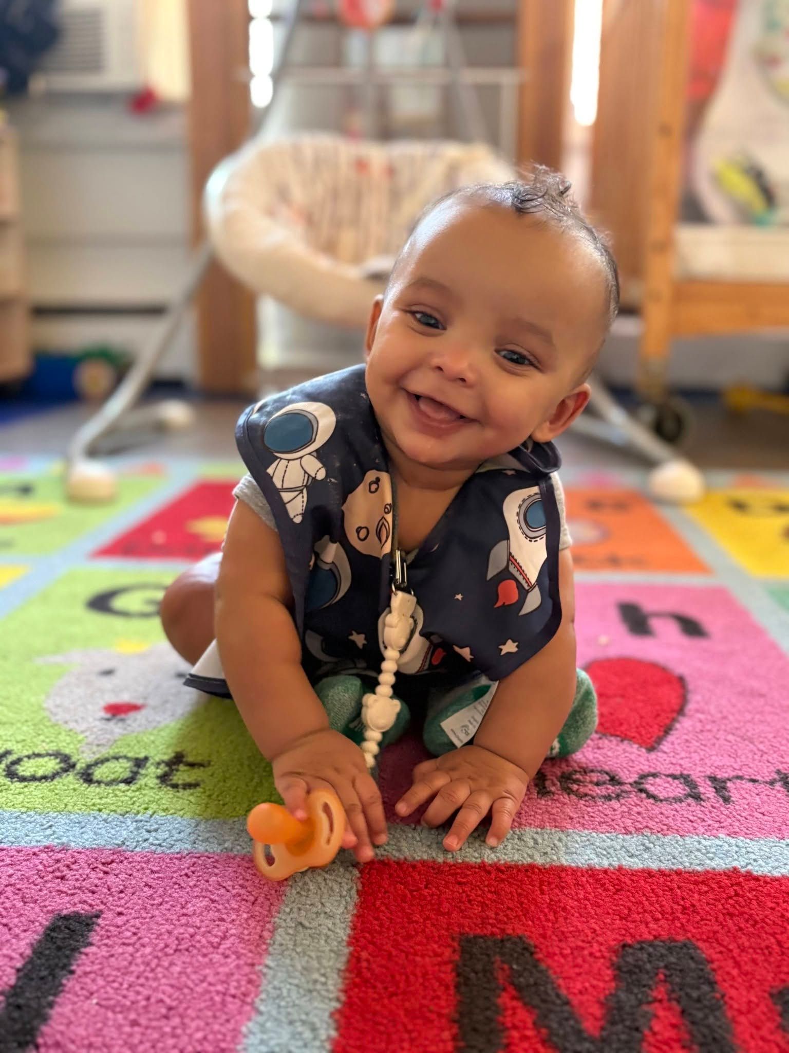 Smiling baby on a colorful play mat, leaning forward and holding a toy in a bright room