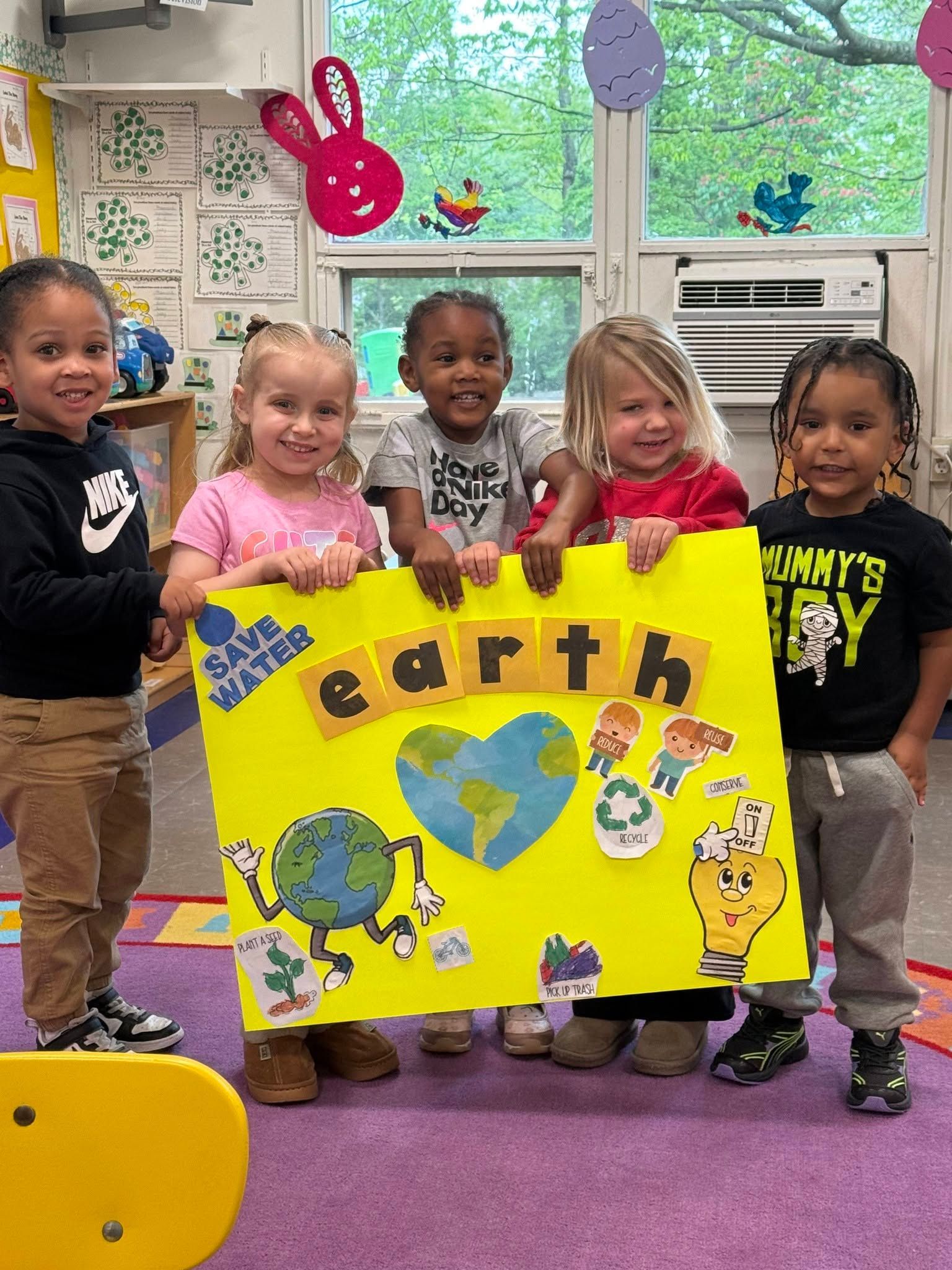 Five children hold a bright yellow Earth poster in a classroom, smiling in front of a window.