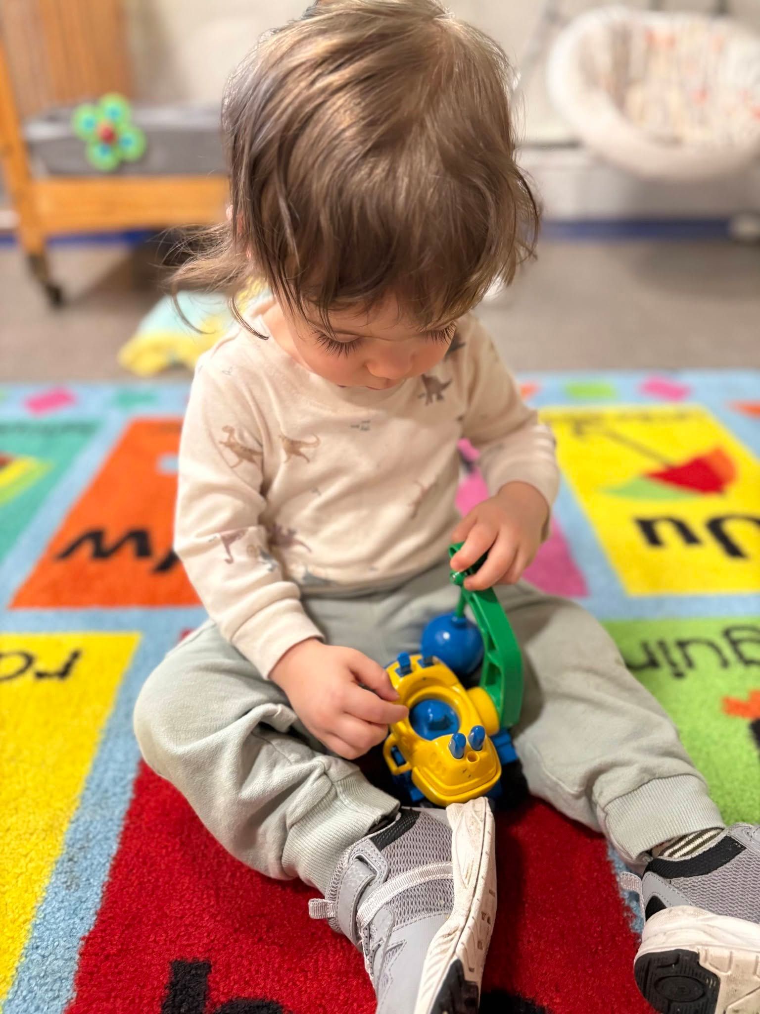 Toddler sitting on a colorful rug, playing with a yellow-and-blue toy car.