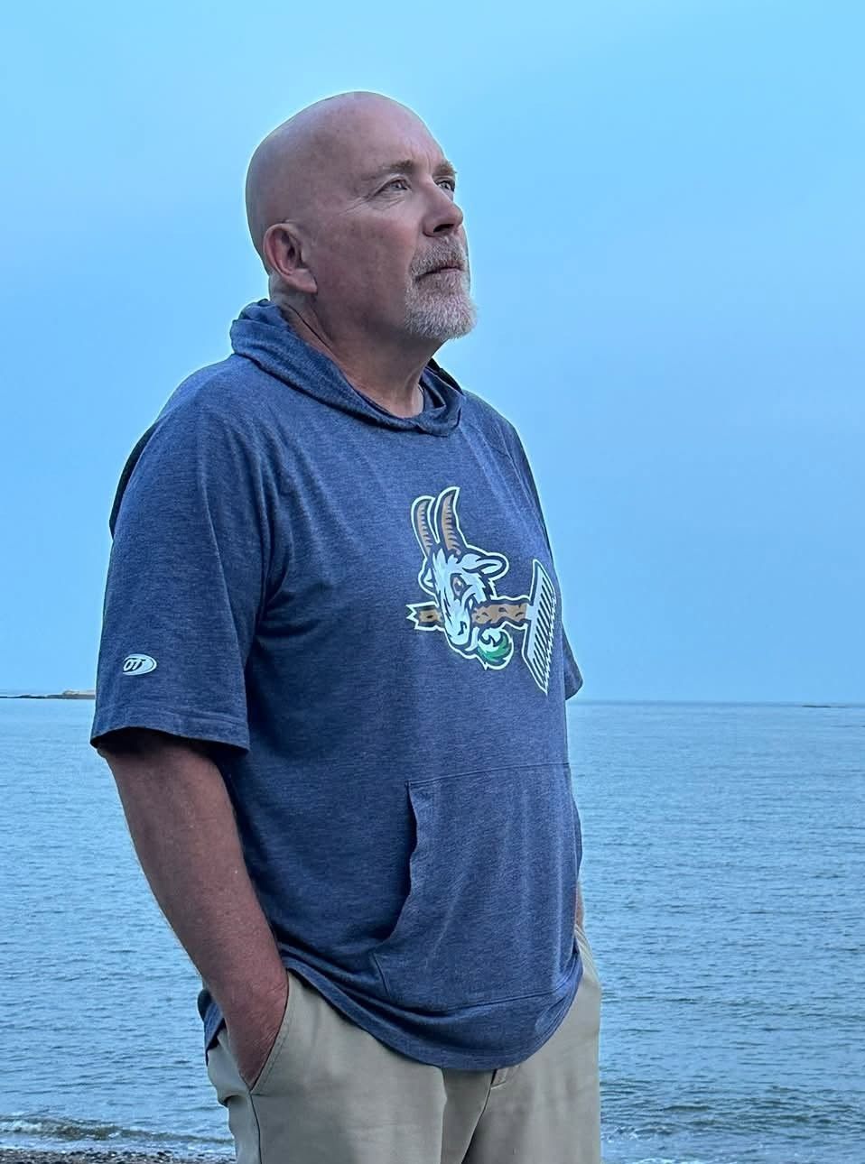 Football coach, arms crossed, on the field. Blue shirt, serious expression, players in background.
