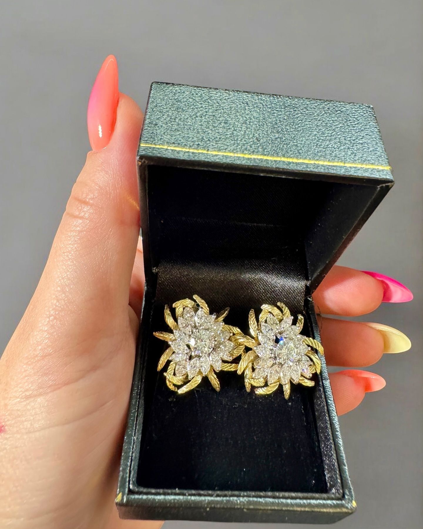 A hand holding an open dark jewelry box containing two decorative, triangular-shaped gold and diamond-encrusted earrings.