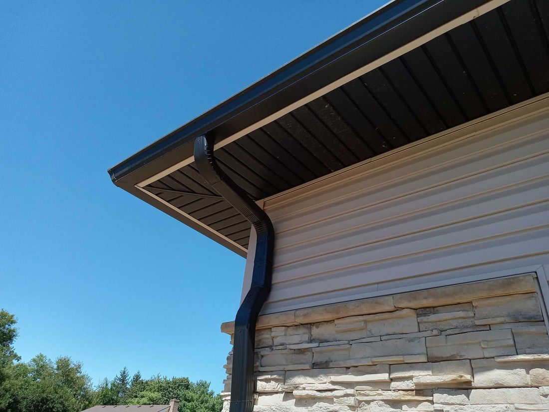 Black gutters and trim on a beige house with stone accents, set against a bright blue sky.