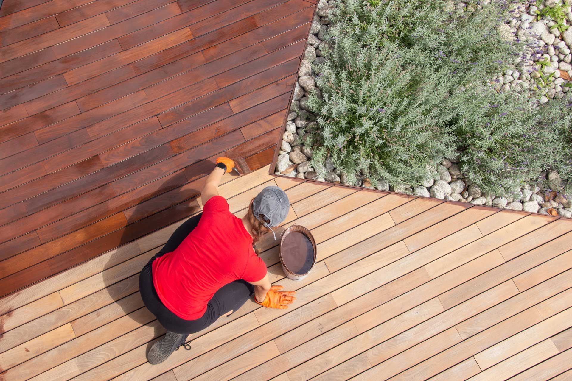 Person applying stain to a wooden deck with a brush, next to a garden.
