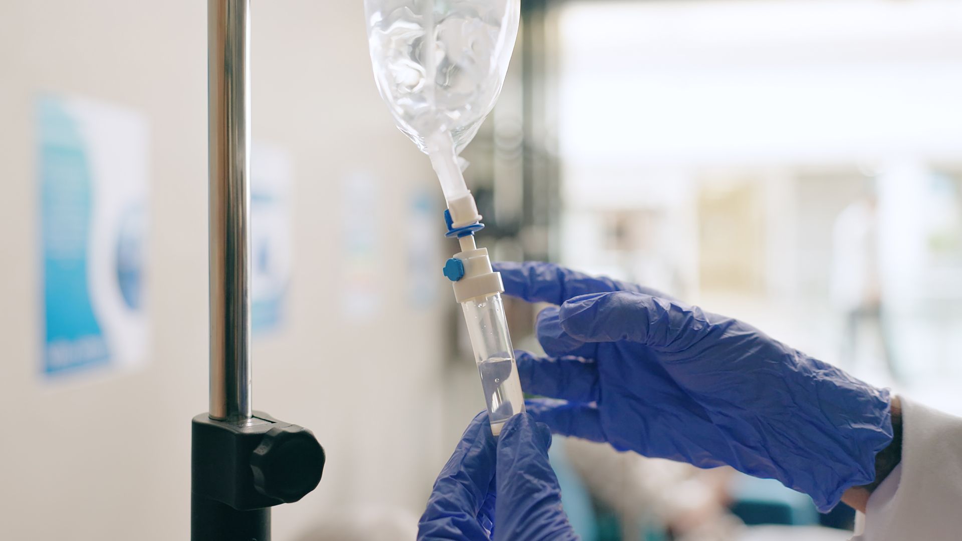 A gloved hand adjusts the flow of a clear intravenous drip bag attached to a metal stand in a clinical setting.