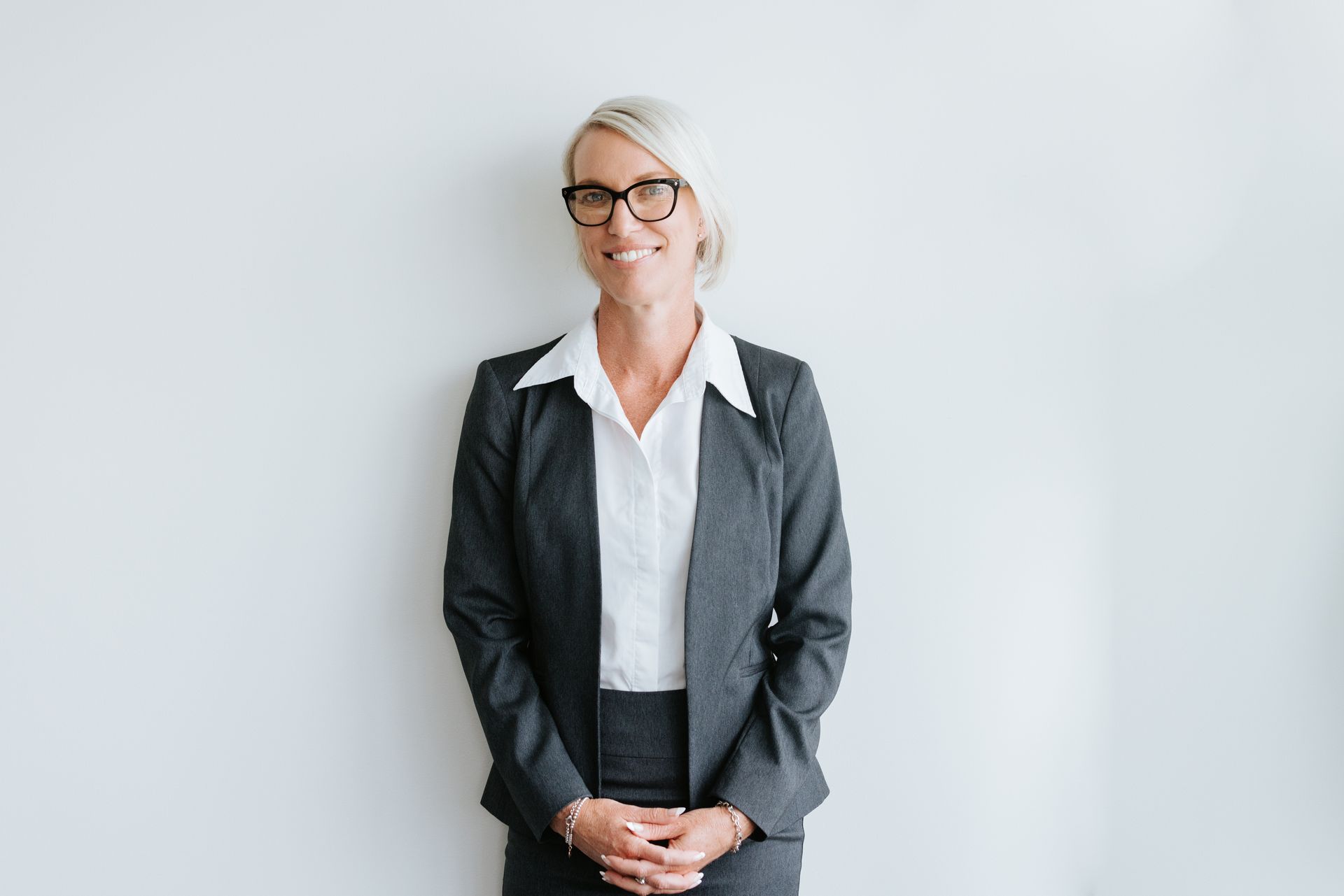 Woman in a dark suit, glasses, and white shirt, smiling, hands clasped, against a white wall.