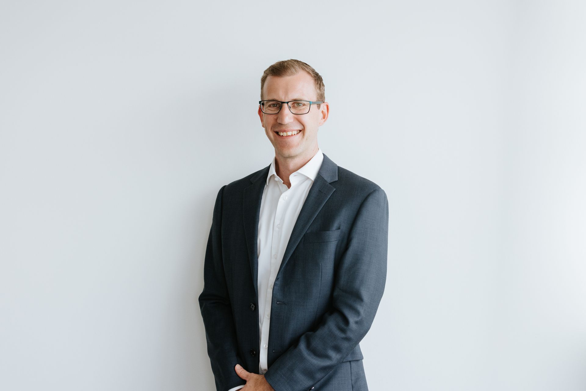 Man in a suit and glasses smiles against a plain white wall.