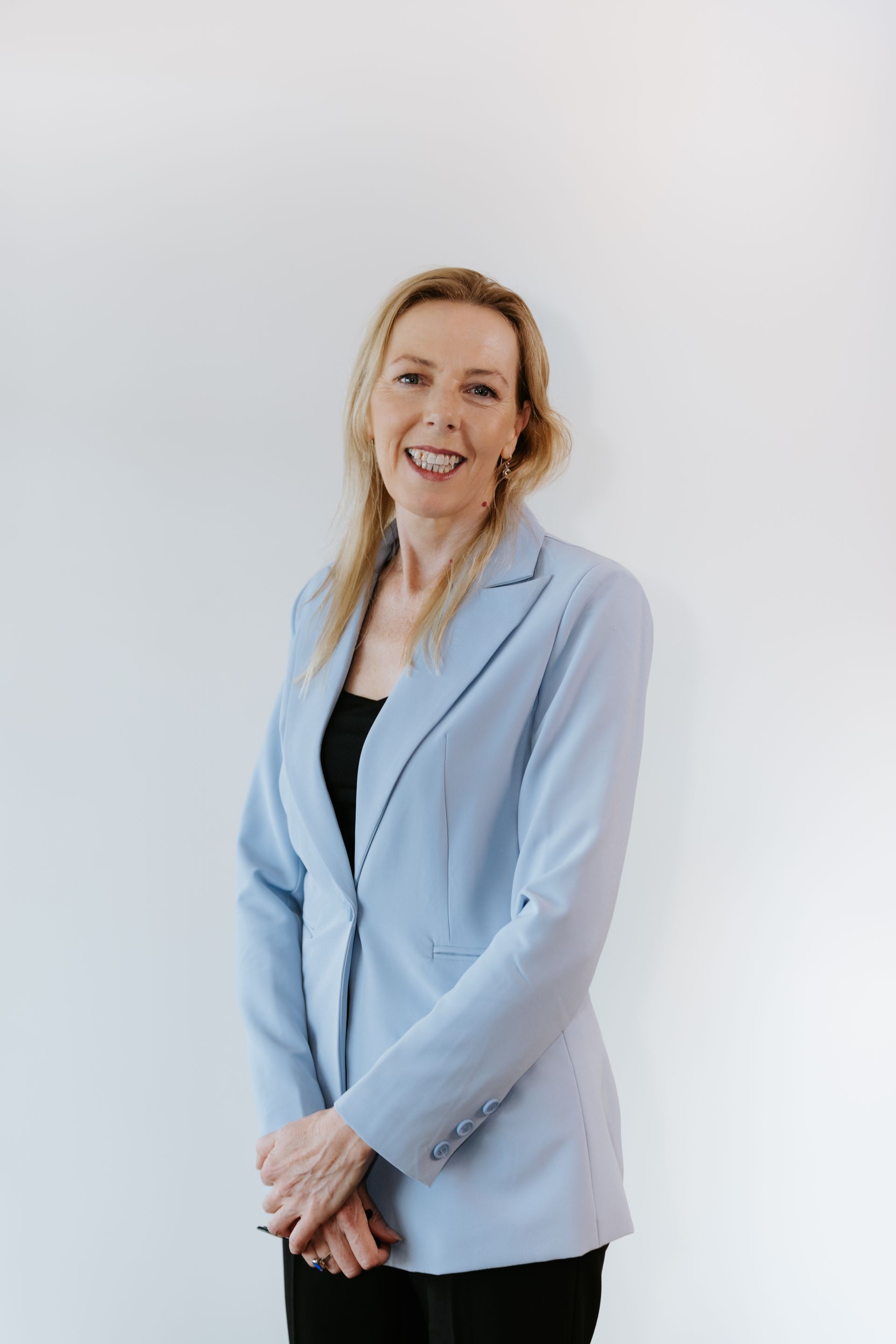 Woman in a light blue blazer smiles at the camera in front of a white wall.