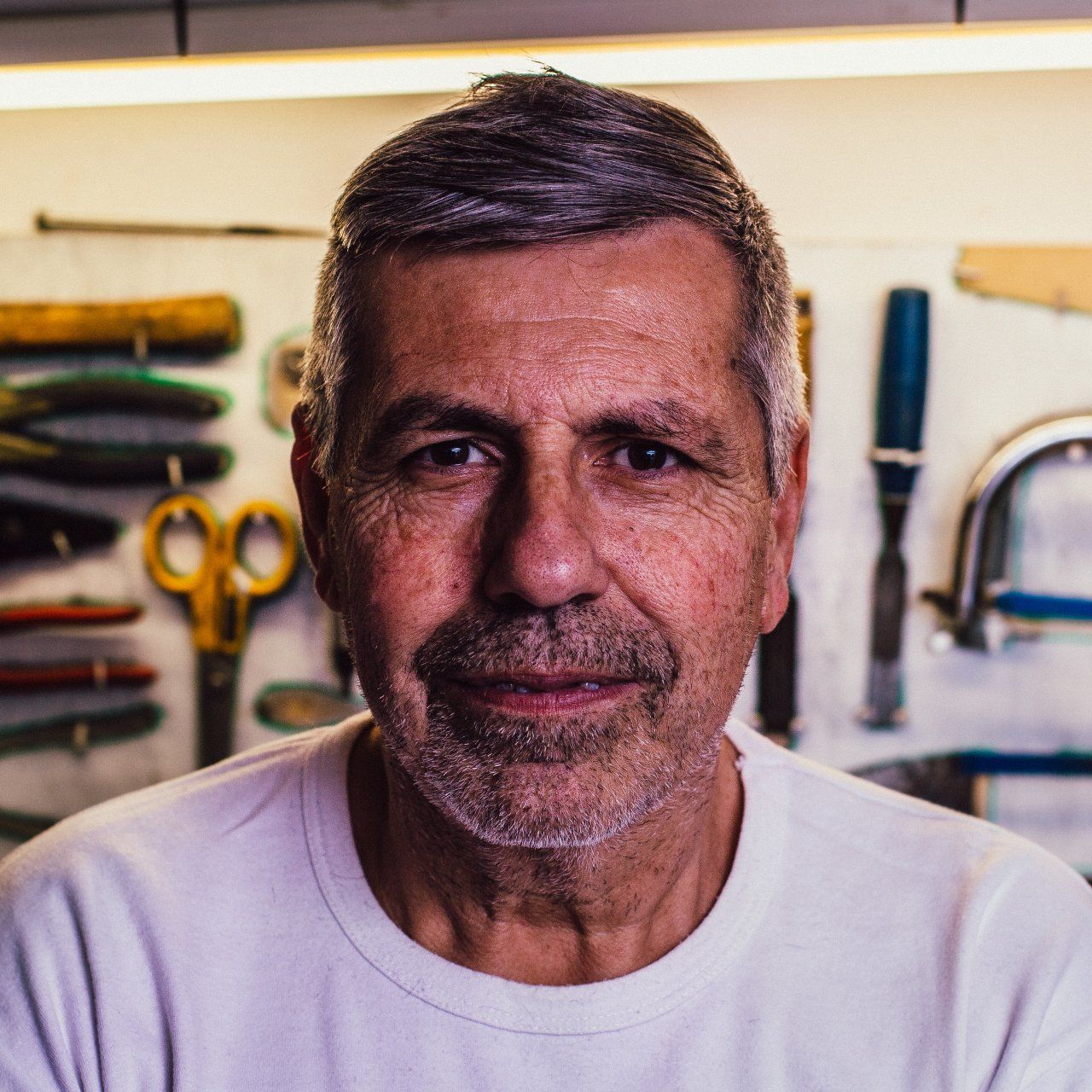 Man with graying hair and beard smiles in a workshop with tools on the wall.