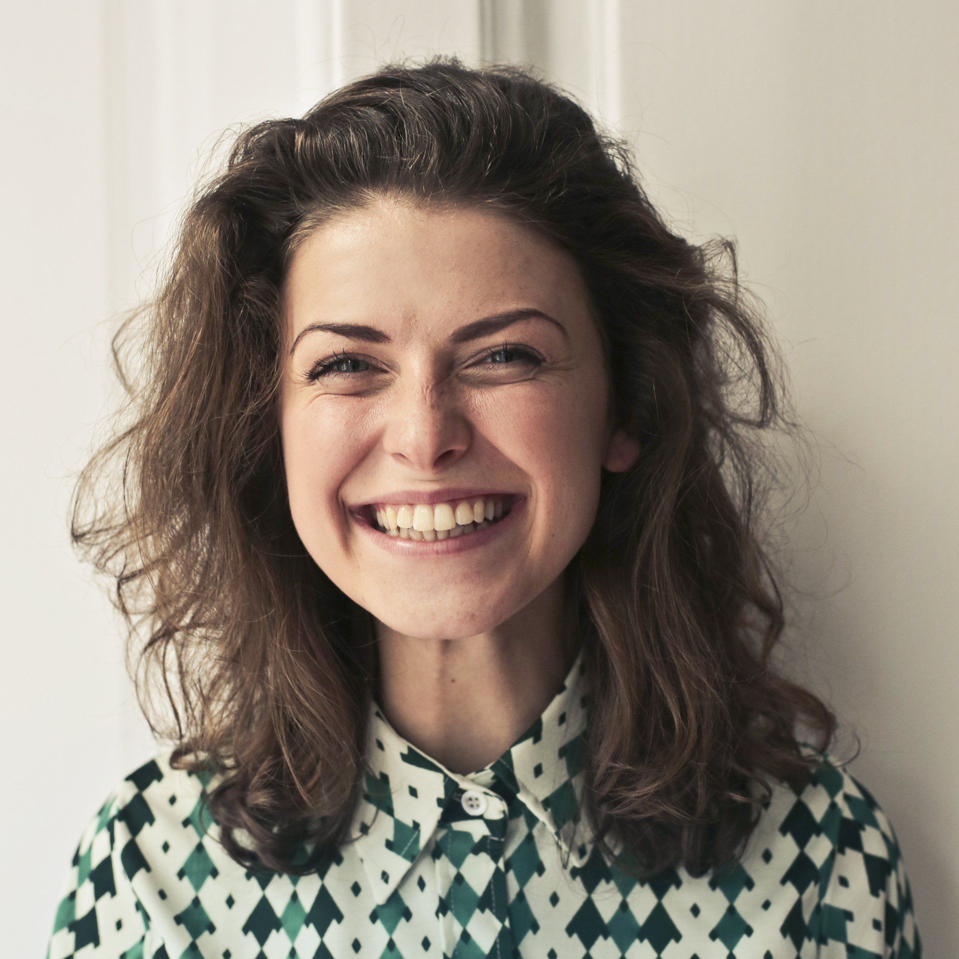 Woman with brown hair smiling broadly, wearing a patterned shirt, indoors.