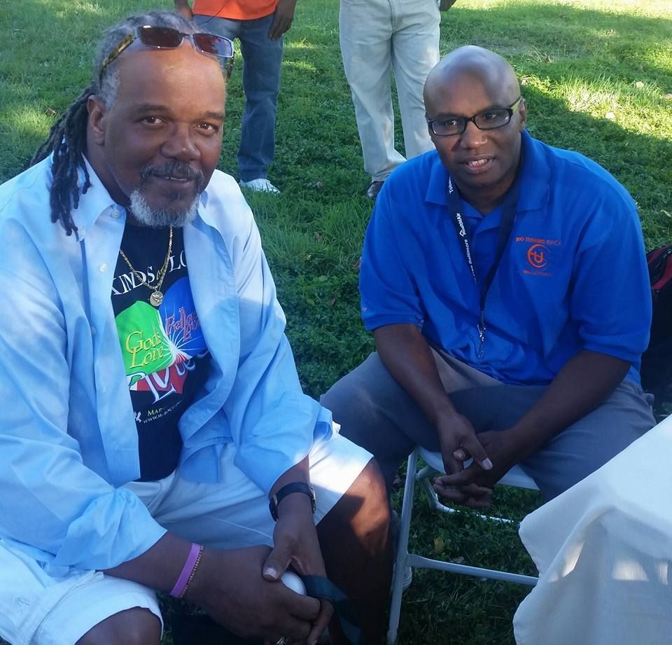 A man wearing a blue shirt with the letter l on it sits next to another man
