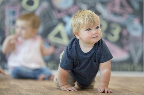 Two children crawling on a wood floor in front of a chalkboard decorated with colorful chalk drawings.