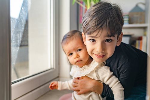 A smiling older child gently hugs a younger child in front of a window.