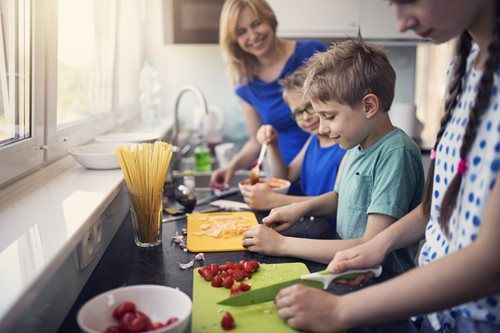 A family prepares a meal together in a sunny kitchen, chopping vegetables on cutting boards beside a jar of pasta.