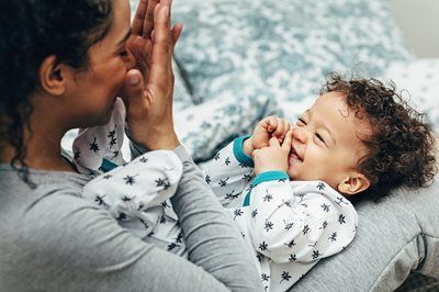 A smiling baby in a patterned onesie plays patty-cake with an adult while lying on their lap.