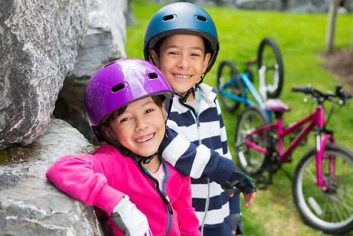A smiling child in a purple helmet and pink jacket stands next to another child in a blue helmet and striped shirt.