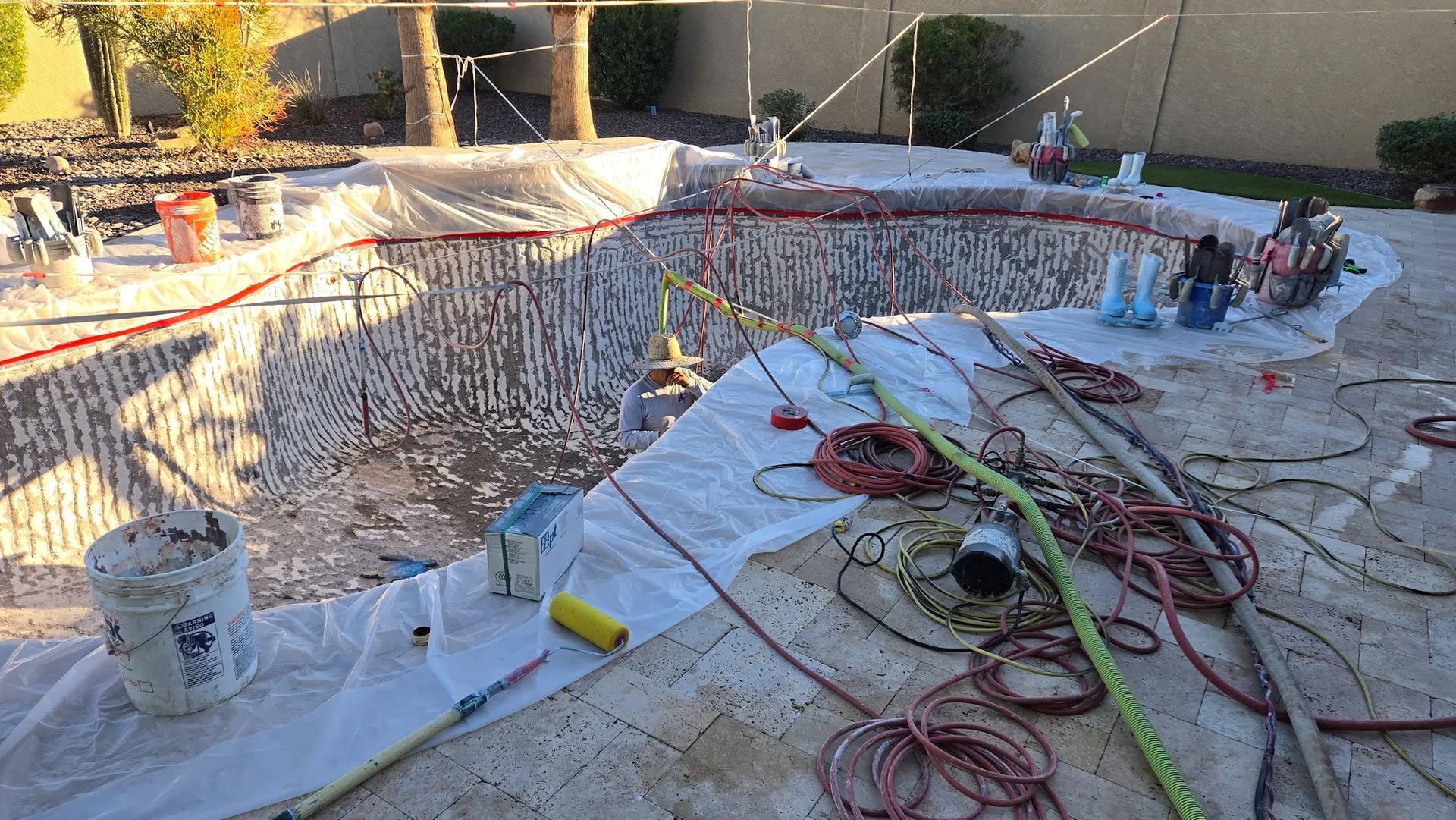 Backyard pool under construction, with workers, hoses, tools, and exposed pool walls covered in netting