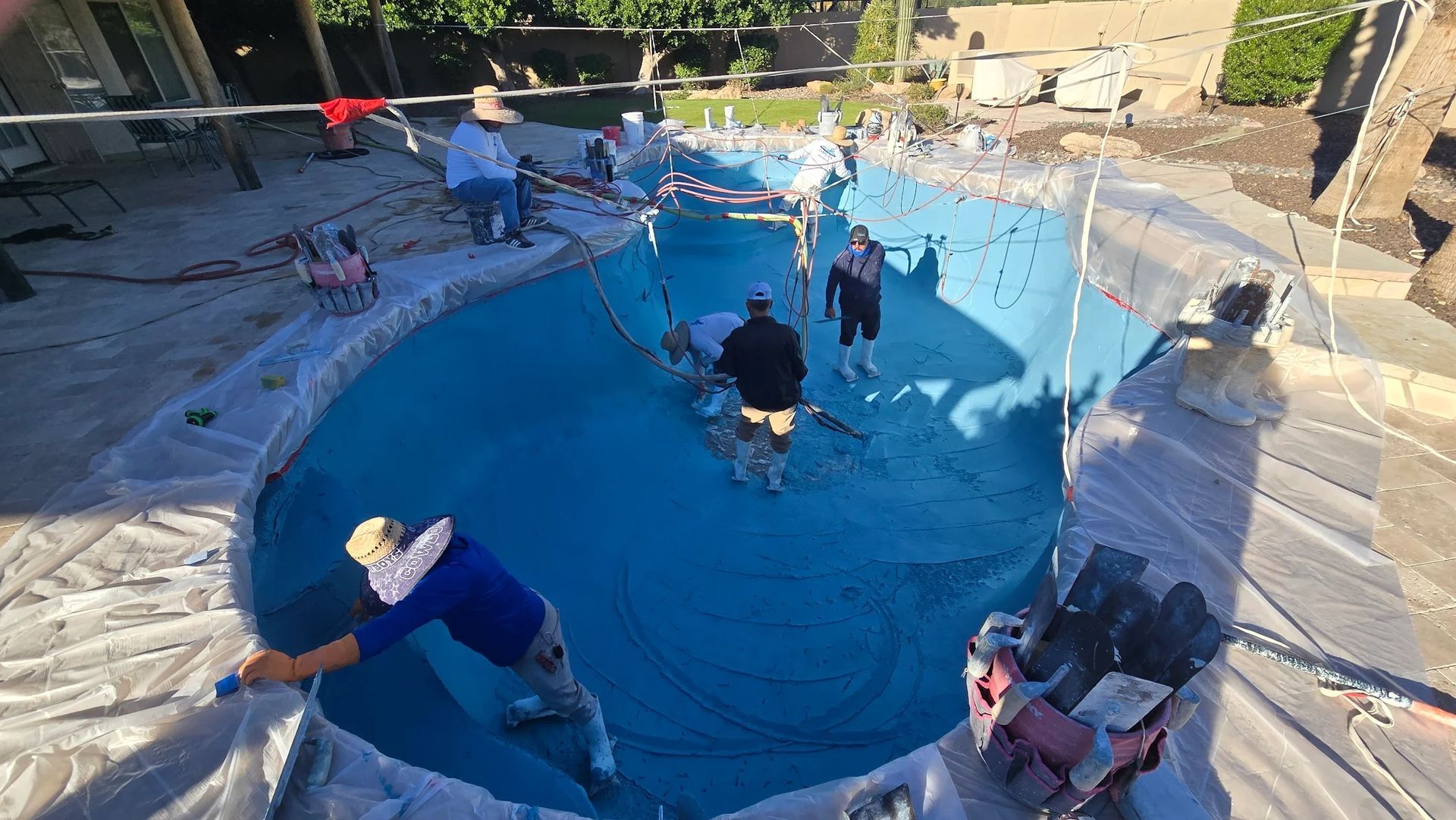 Workers cleaning an empty backyard pool with blue liner and scattered tools in bright sunlight