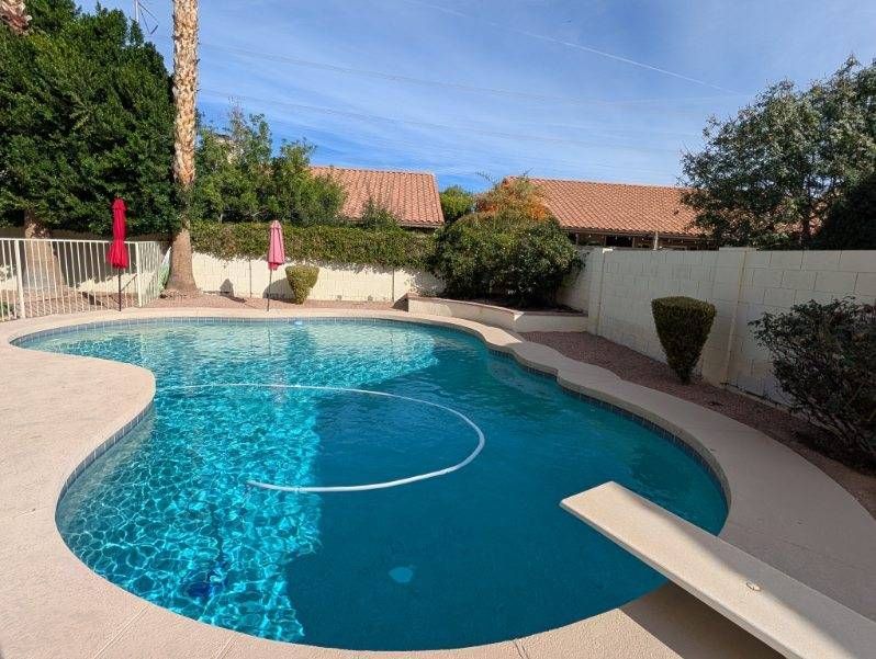 Backyard swimming pool with blue water, diving board, lounge chairs, and tiled roof houses in the background
