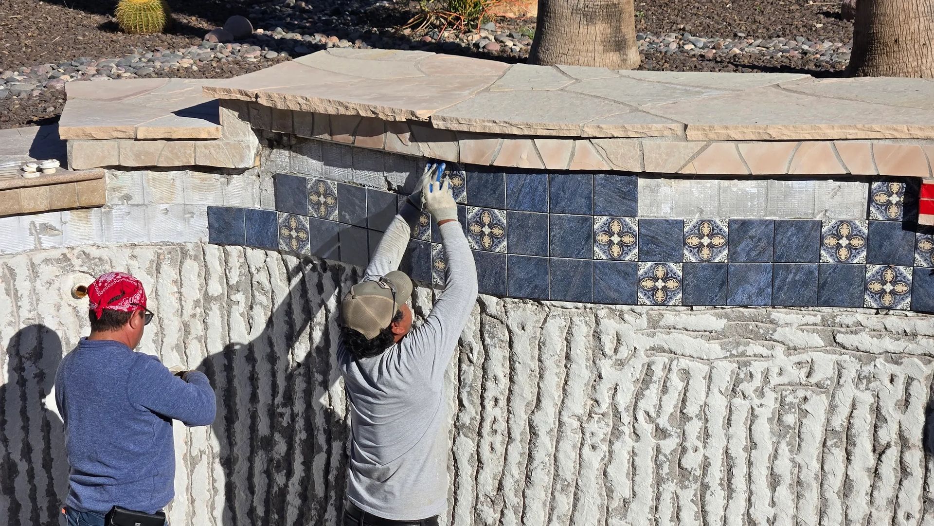 Two people installing a blue and white mosaic tile wall outdoors