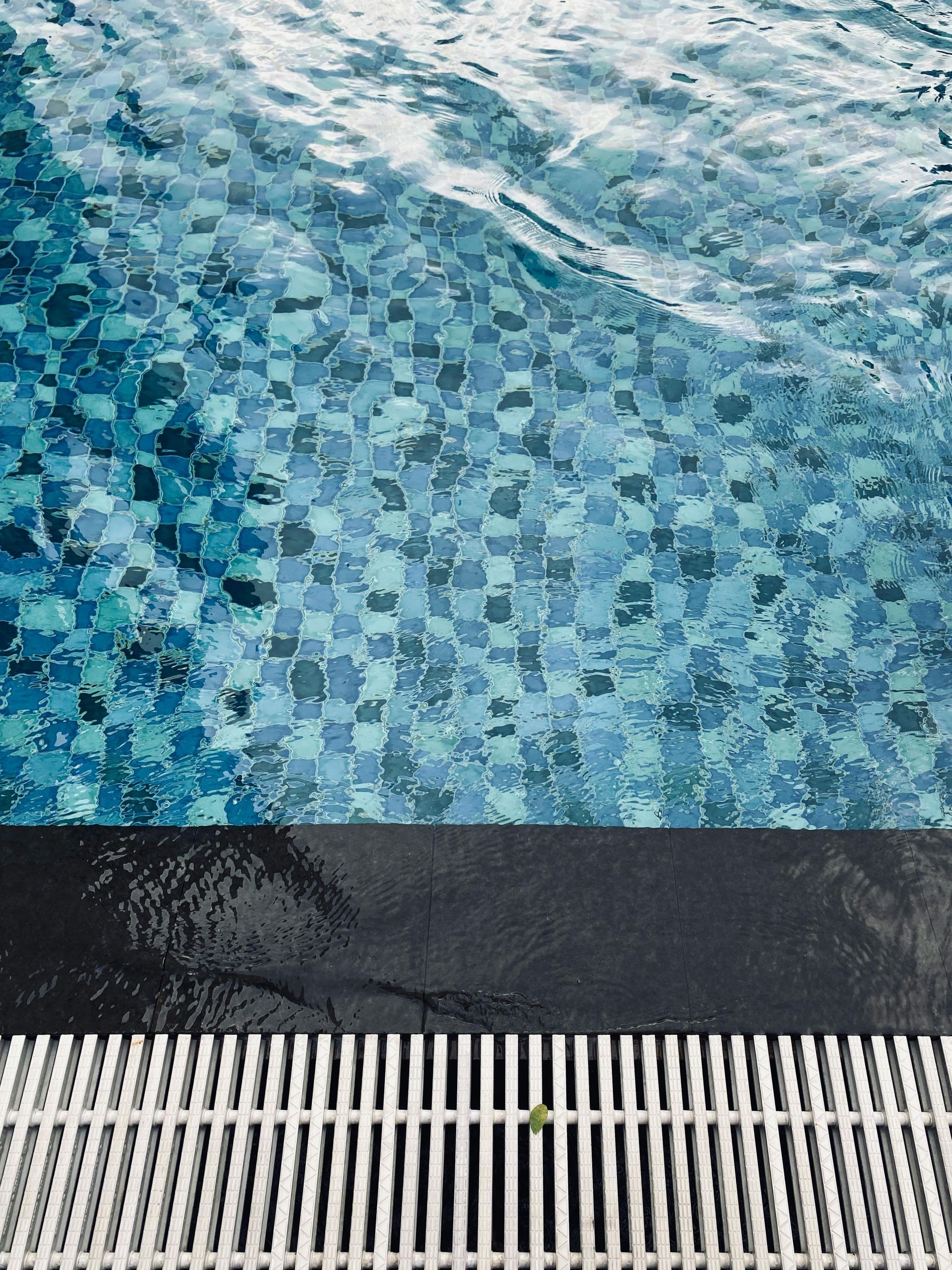 Blue mosaic pool water beside a dark pool edge and white drainage grate