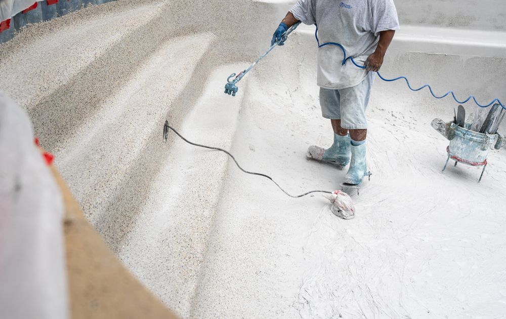 Worker spray-painting a white floor with a hose and paint sprayer in a bright room