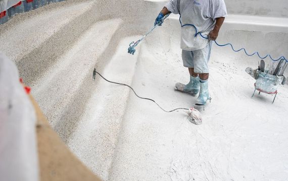 Worker spray-painting a white floor with a hose and paint sprayer in a bright room