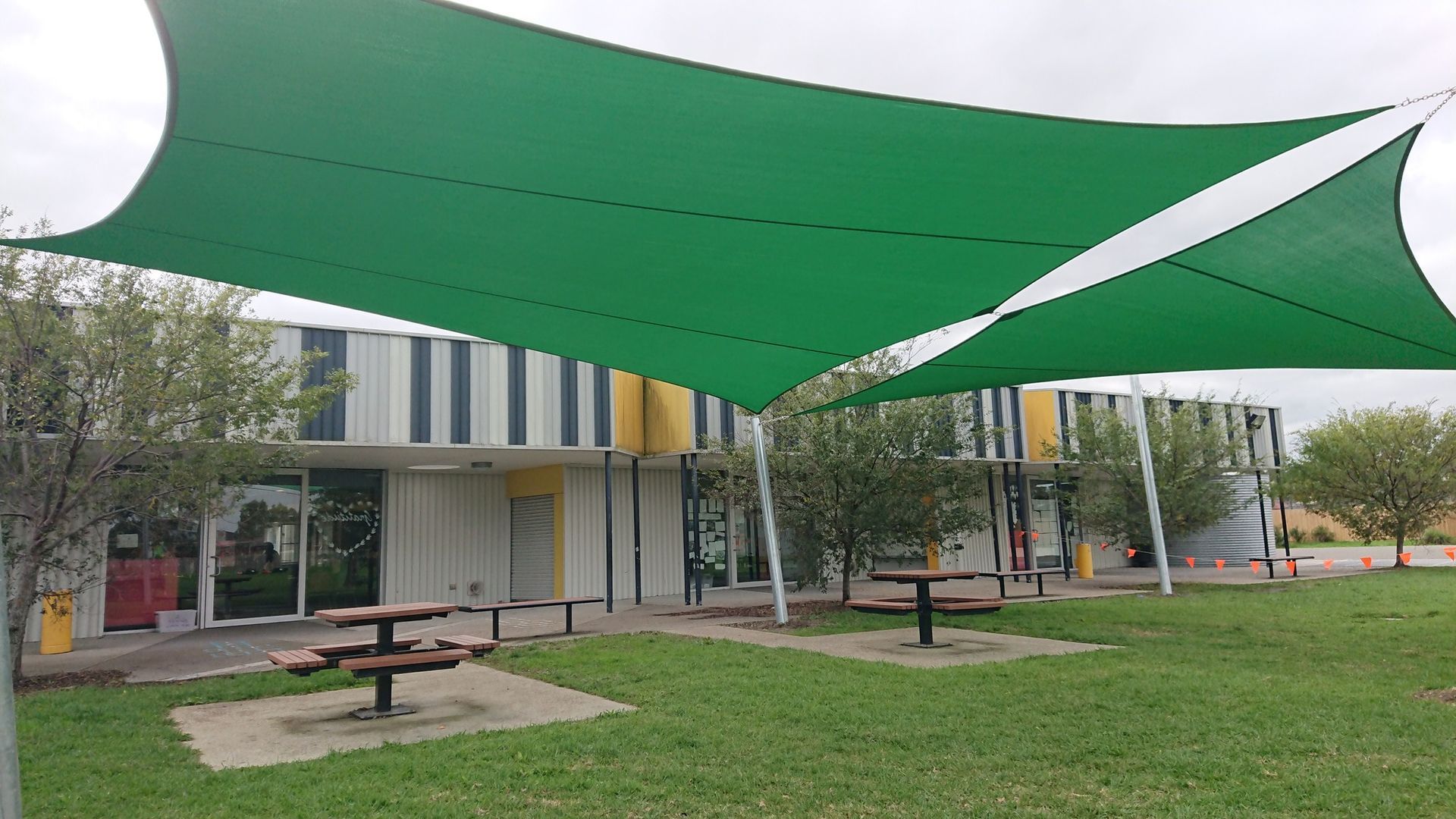 Shade sail installation at St John’s Regional College, 5/11 Caroline St, Dandenong VIC 3175—featuring linked green rectangular hyperbolic canopies supported by steel posts, providing sun protection over picnic tables on concrete pads in a landscaped courtyard beside a colourful striped school building