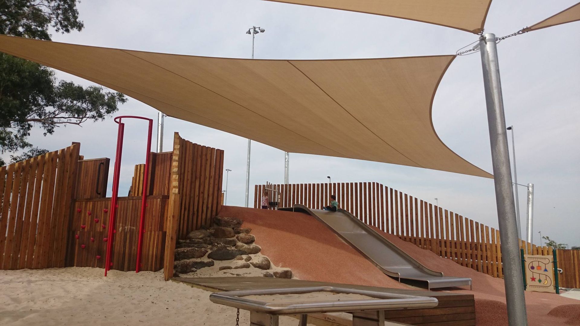 Shade sail installation at Livvi’s Place Casey, Boland Dr, Lyndhurst VIC 3975—featuring large beige canopies supported by steel posts, providing sun protection over an inclusive playground with a climbing wall, metal slide, sandpit, and natural rock landscaping designed for accessibility and family-friendly use