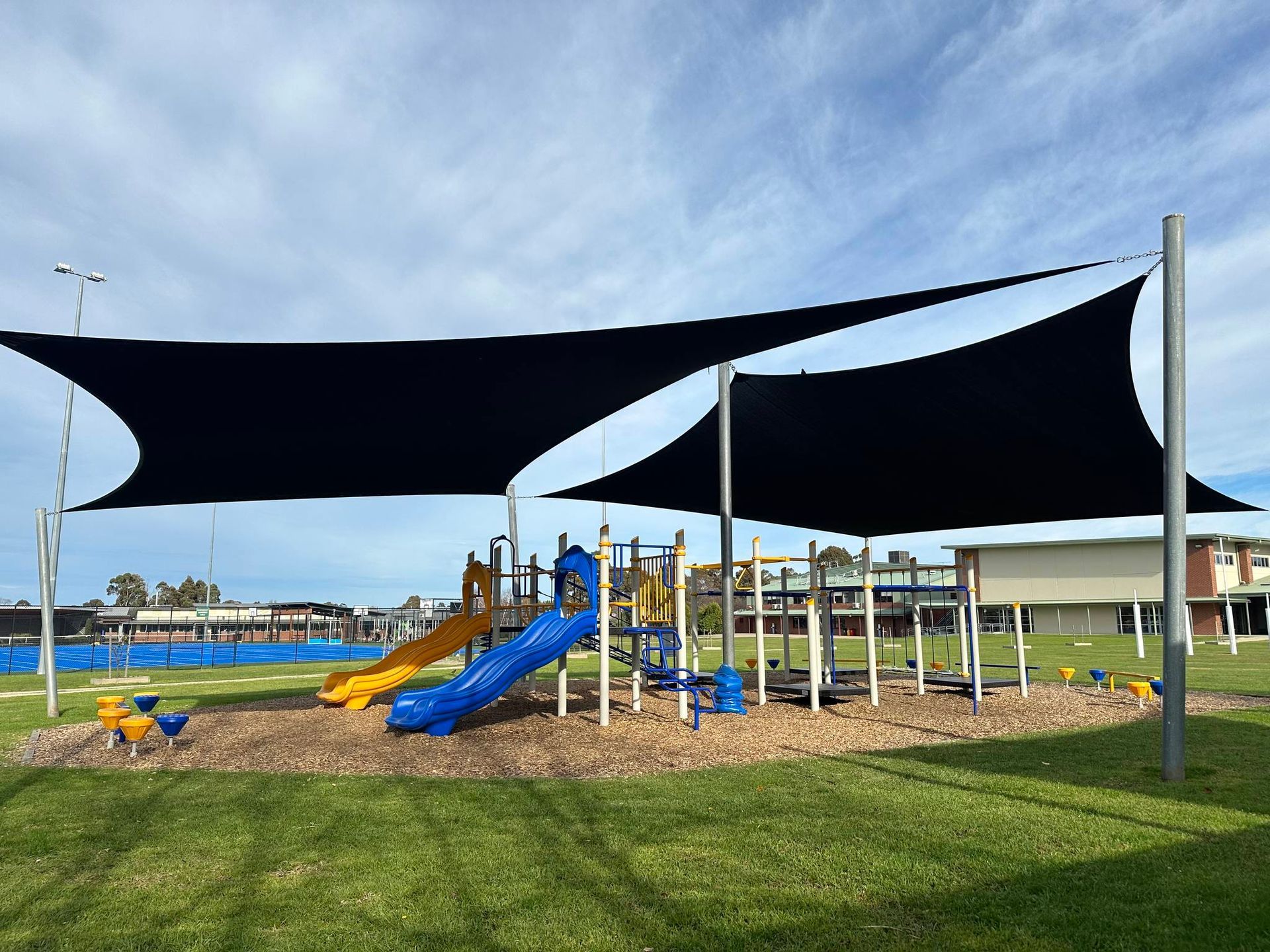 Commercial-sized black shade sails covering entire playground at Beacon Hills College in Pakenham, featuring triangular canopies supported by tall steel posts, shading multiple slides and climbing structures for student sun protection in landscaped school setting