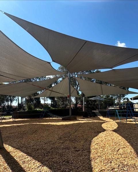 Custom commercial shade sail installation at Fairhaven Family and Children’s Centre, 52 Fairhaven Blvd, Cranbourne West VIC 3977—featuring multiple beige triangular canopies converging at central points, supported by tall steel posts and providing shaded coverage over a woodchip playground with swings and climbing structures in a landscaped early learning environment
