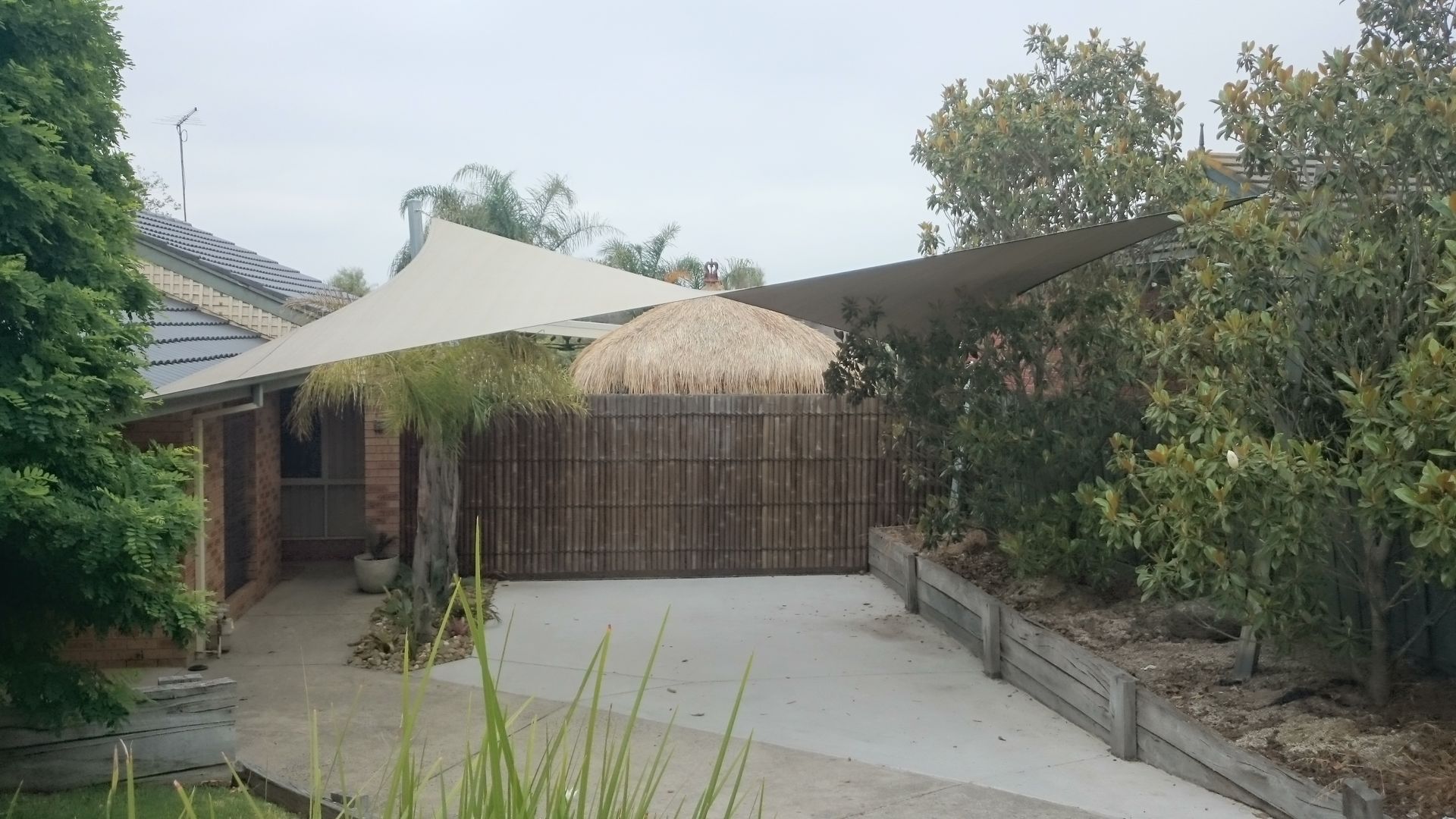 Modern shade sail installation for a residential carport in Berwick 3806, featuring hyperbolic rectangular shade sail tensioned over a concrete driveway to enhance the property’s aesthetics and functionality. Anchored to the house and perimeter posts, the design complements the natural surroundings with greenery, bamboo fencing, and a thatched-roof feature in the background. Professionally installed by a licensed Melbourne shade sail expert