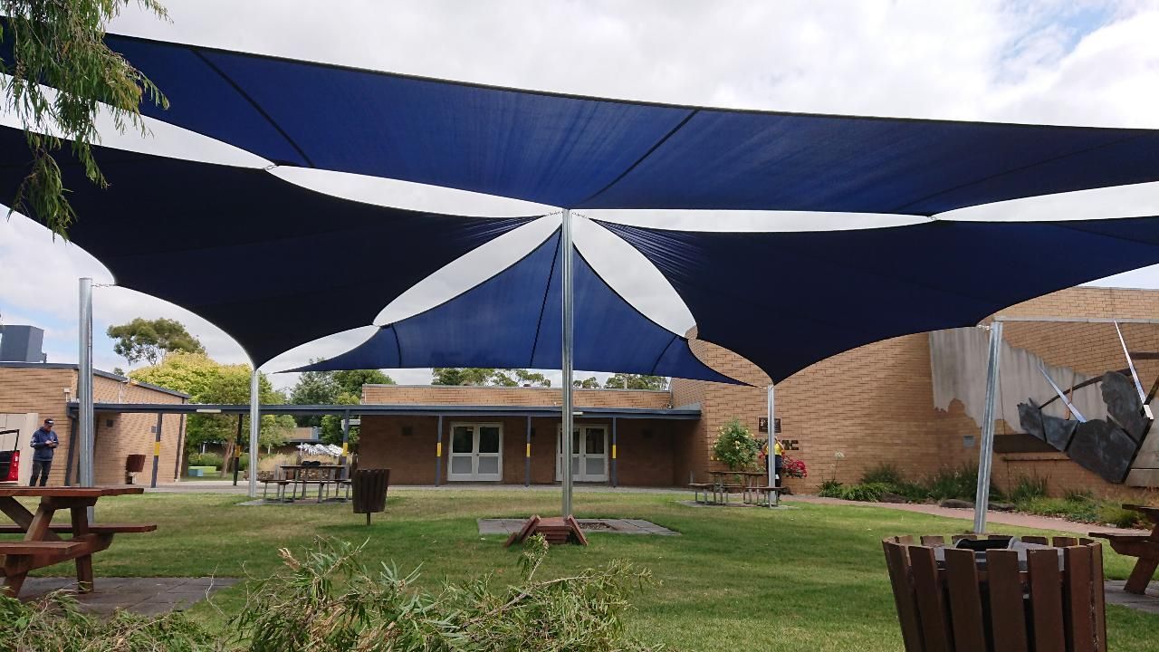 Shade sail installation at Lynbrook Primary School, 37 Paterson Dr, Lynbrook VIC 3975—featuring a blue and white canopy supported by metal posts, providing sun protection over outdoor picnic tables near a brick school building in a grassy courtyard