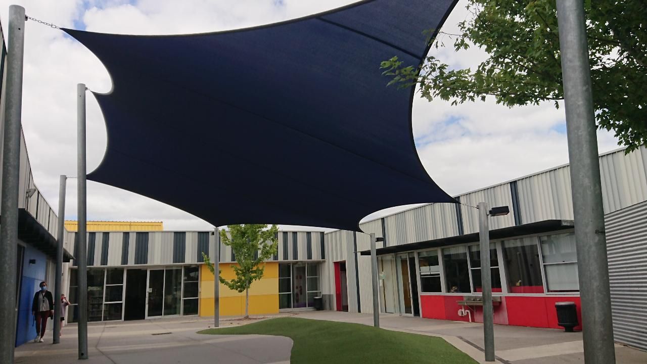 Modern shade sail installation at St Francis de Sales Primary School, 122 Paterson Dr, Lynbrook VIC 3975—featuring a large, dark blue canopy supported by steel posts, shading a colourful outdoor area surrounded by contemporary school buildings and green landscaping