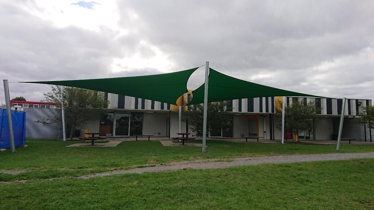 Shade sail installation at St John’s Regional College, 5/11 Caroline St, Dandenong VIC 3175—featuring linked green rectangular hyperbolic canopies supported by steel posts, providing sun protection over picnic tables on concrete pads in a landscaped courtyard beside a colourful striped school building