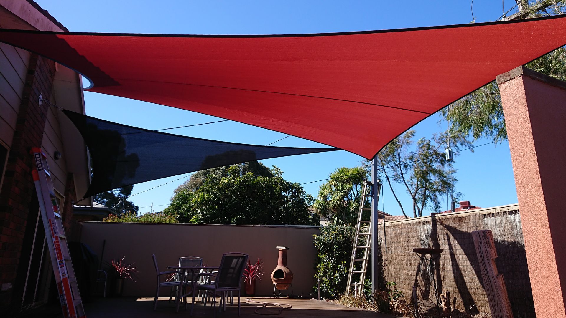Custom red and black shade sail installation over timber decking at a private home in Rowville, designed to provide year-round UV and weather protection for outdoor entertaining area with garden surrounds and outdoor furniture