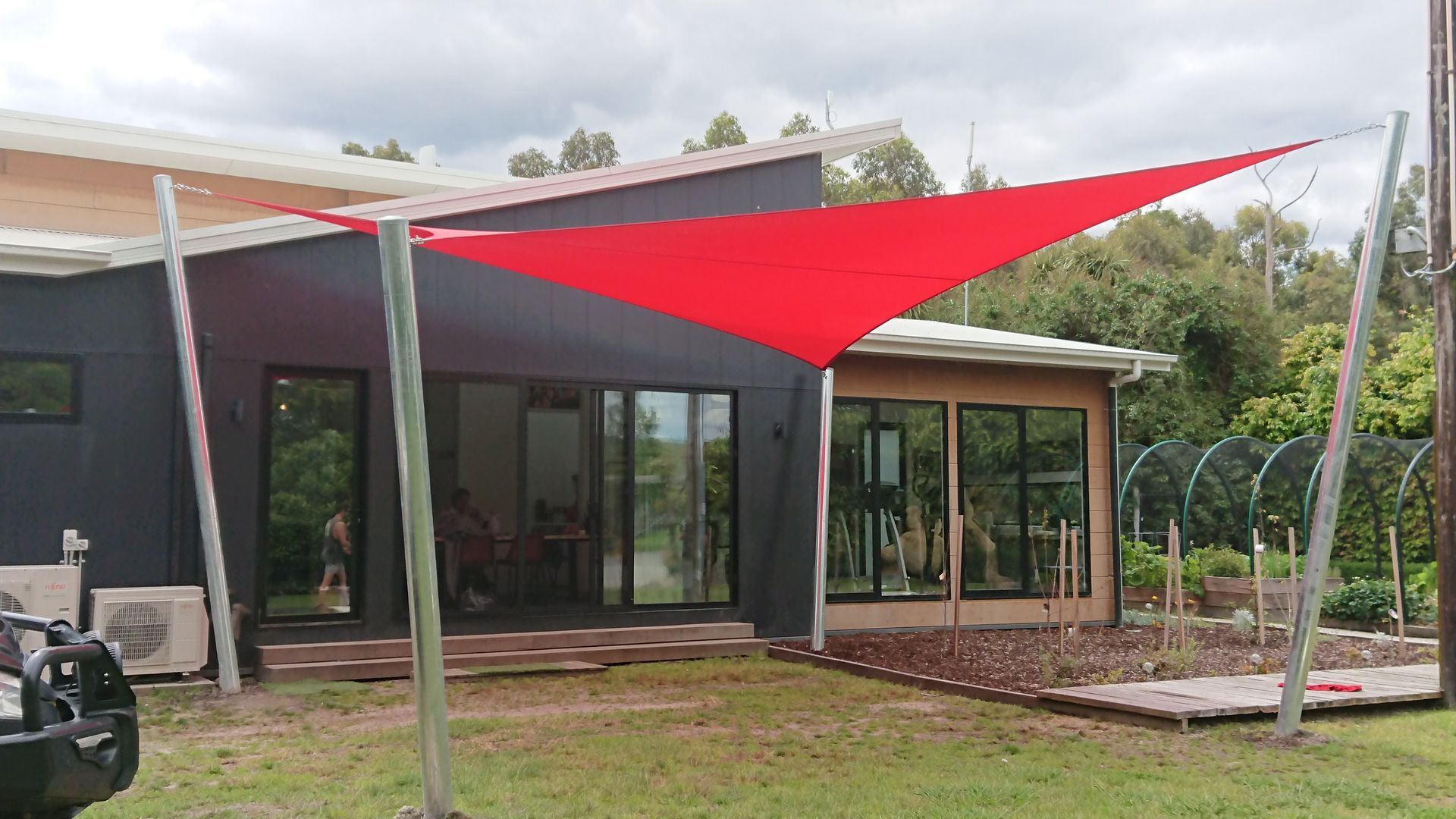 Shade sail installation at a residential property in Labertouche VIC 3816—featuring a bold red rectangular hyperbolic twist canopy supported by steel posts, extending from the modern house façade to create a shaded outdoor area beside a garden with raised beds and a wooden deck, enhancing both functionality and visual appeal