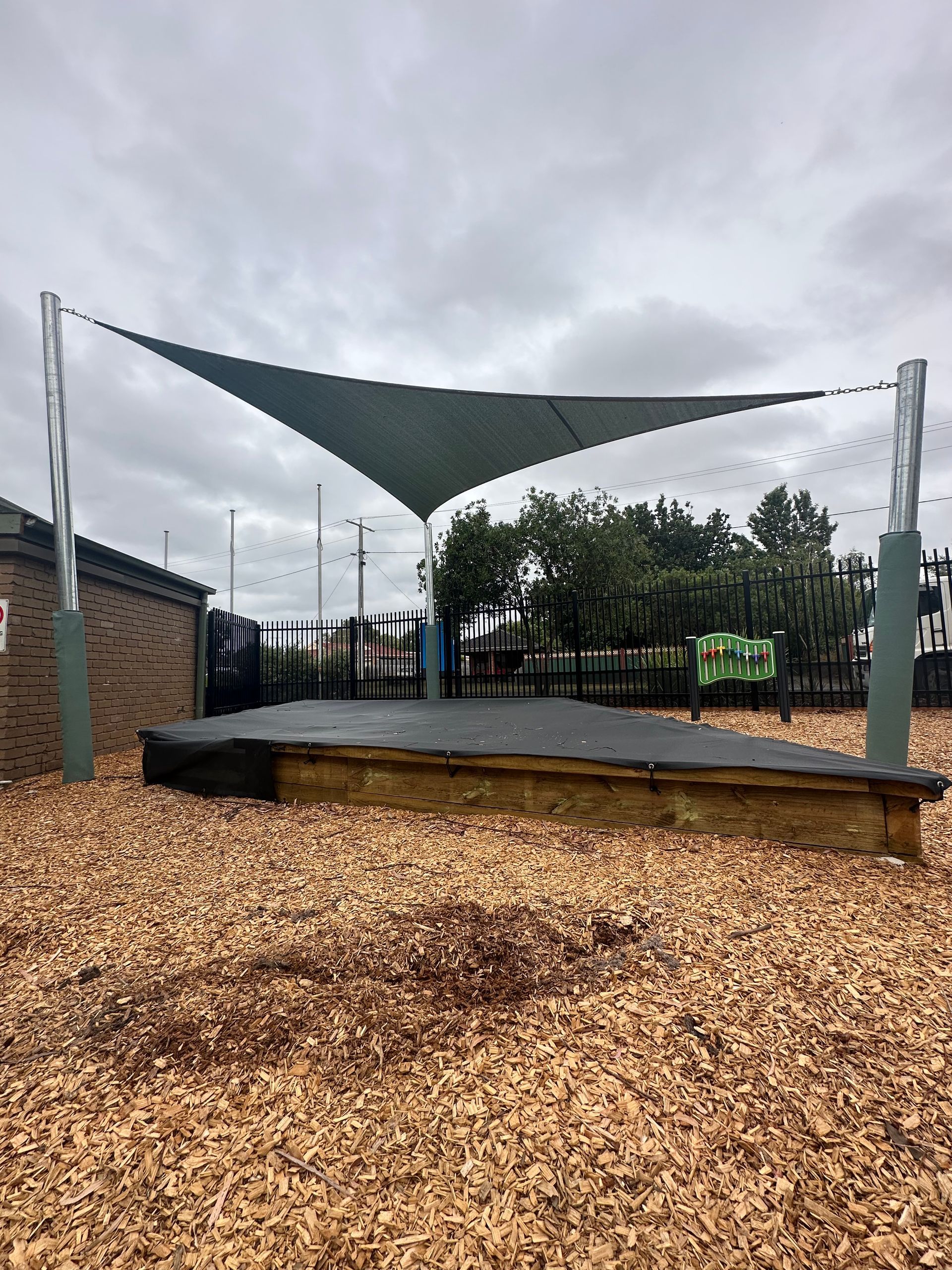Shade sail installation at Blind Bight Community Centre, 42 Anchorage Dr, Blind Bight VIC 3980—featuring a large triangular and wave panel canopy supported by steel posts, providing sun protection over a sandpit area surrounded by wood chips, with a fenced playground and green play equipment 