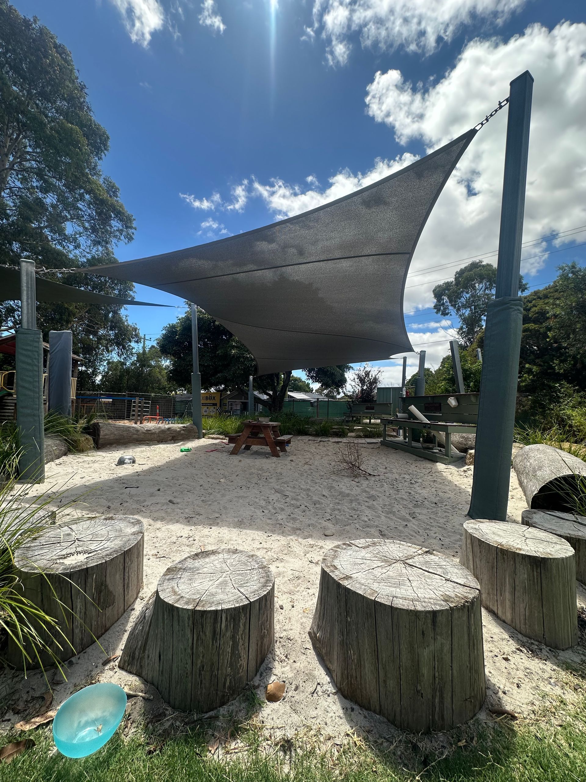 Shade sail installation at Ballam Park Preschool, 26R Naranga Cres, Frankston VIC 3199—featuring large hyperbolic twist and wave panel commercial shade sail fabric canopies supported by tall metal posts, providing shade over a natural outdoor play area with a sandpit, swing set, tree stumps, and surrounding greenery