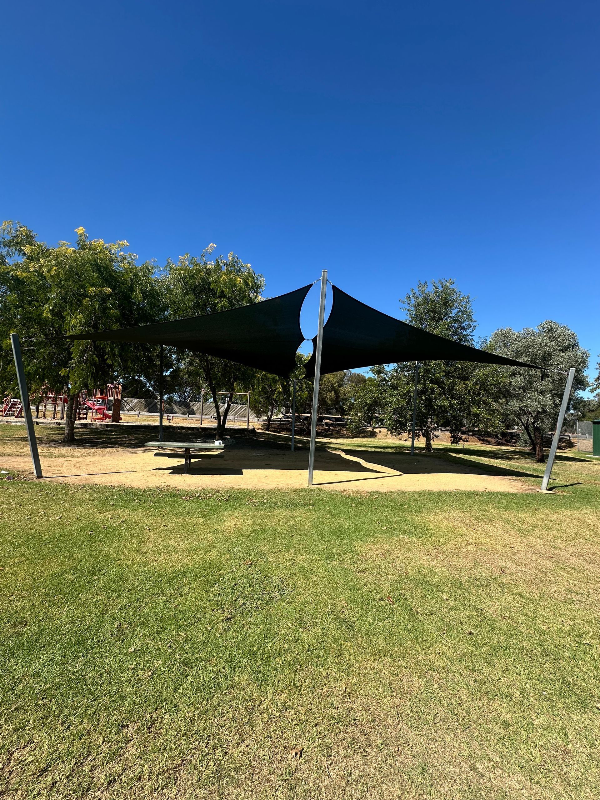 Custom commercial shade sail installation at Sydney Pargeter Reserve Playground, Sydney Pargeter Recreation Area, Kennington Park Dr, Endeavour Hills VIC 3802—featuring two joined square hyperbolic twist canopies over each football oval, tensioned between steel posts to create dynamic architectural shade for public seating areas, with fencing, trees, and recreational facilities in the background