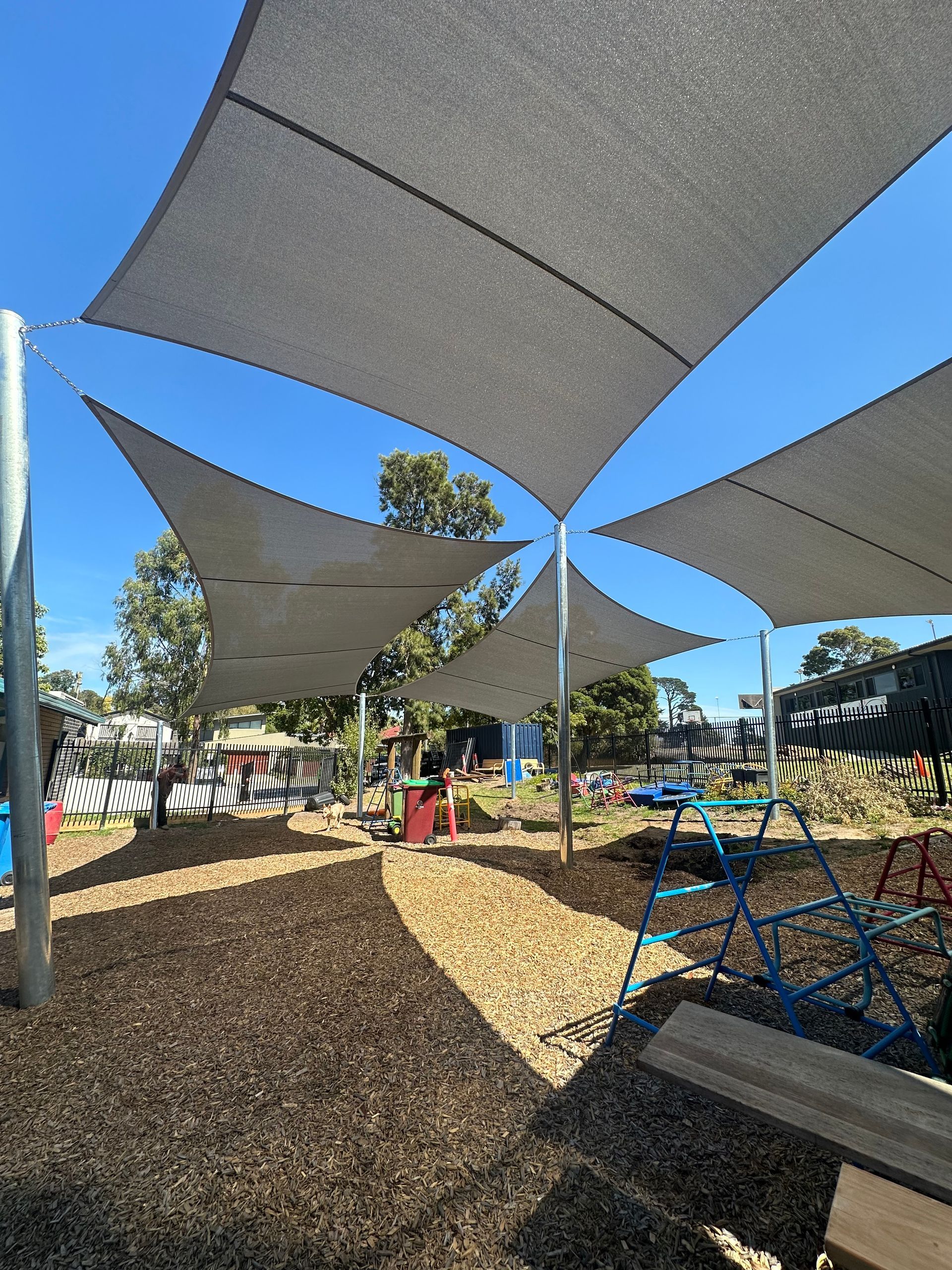 Shade sail installation at Narre Warren North Kindergarten, 8 Winters Ln, Narre Warren North VIC 3804—featuring intersecting hyperbolic square canopies mounted on tall steel poles, providing shade over soft fall play area, playground with climbing equipment and natural surroundings in a secure outdoor setting for children 