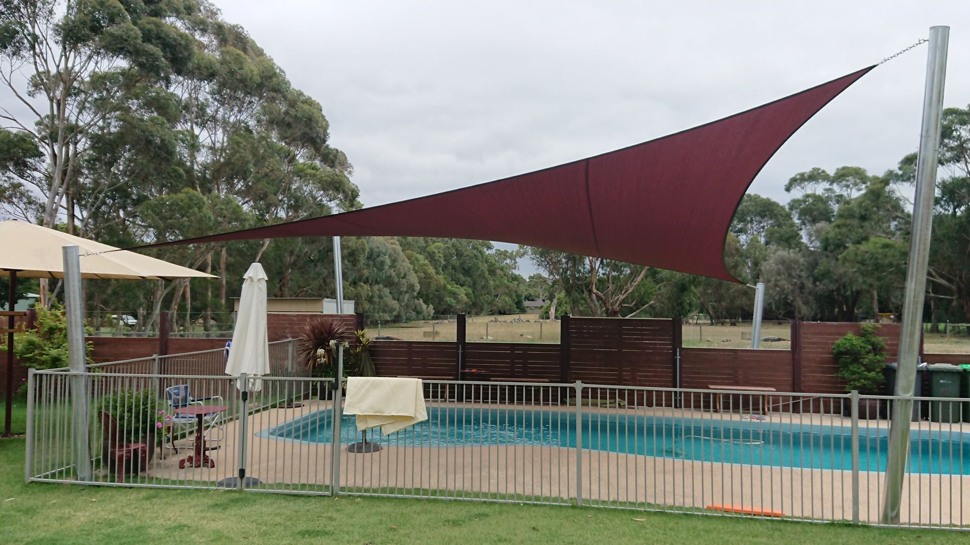 Dark maroon Hyperbolic shade sail installed over residential swimming pool in Pakenham, supported by galvanised metal posts and designed to protect against UV rays while enhancing privacy and outdoor comfort in a landscaped backyard setting