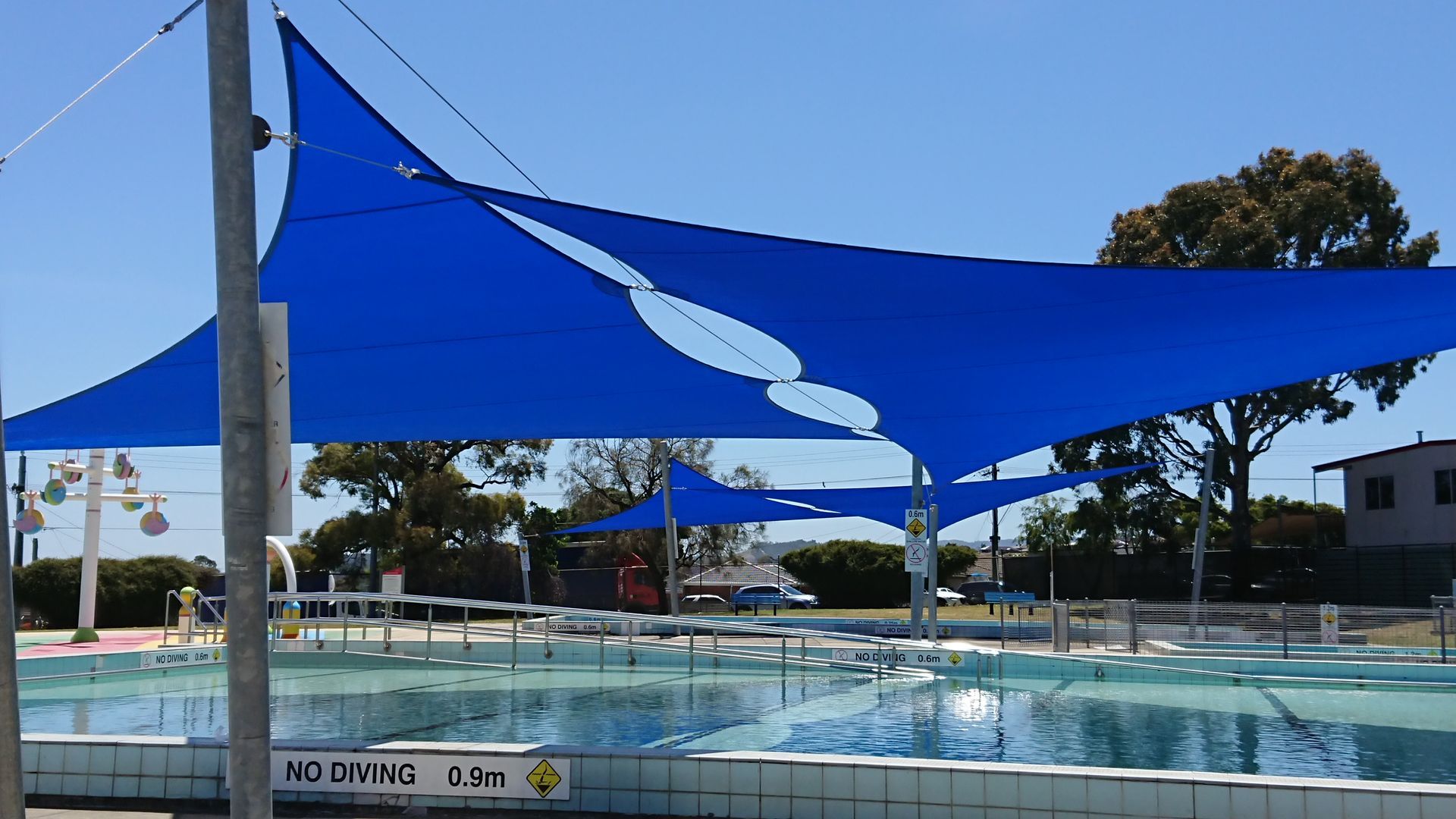 Commercial shade sail installation over outdoor pools at Doveton Pool in the Park, Kidds Rd & Tristania St, 64 Tristania St, Doveton VIC 3177—featuring high-tension blue fabric canopies supported by galvanized steel posts, delivering reliable UV protection and safety over children’s swimming areas, with surrounding secure fencing, signage, and adjacent recreational facilities