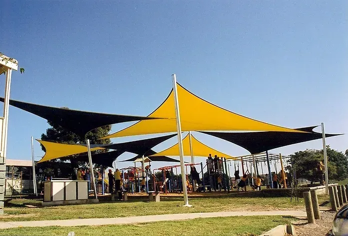 Commercial shade sail installation at Berwick Lodge Primary School, 139-145 Mansfield St, Berwick VIC 3806, featuring yellow and black triangular canopies tensioned over a large playground. Supported by tall white steel posts, the sails provide broad UV protection for various play structures, creating a safe, shaded environment for students. Professionally installed by a licensed Melbourne-based shade sail specialist